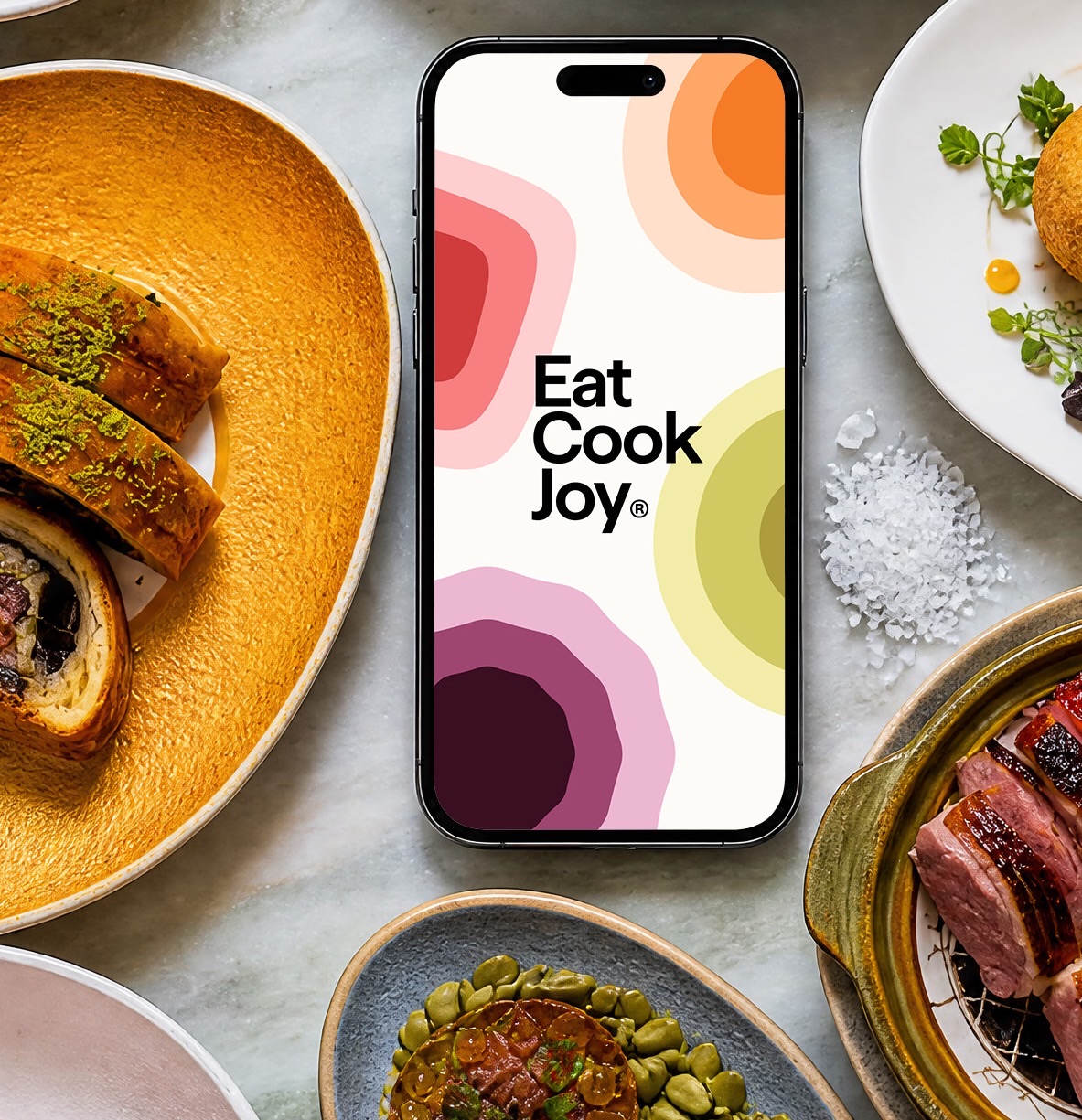 The Eat Cook Joy app on a phone, surrounded by plates of food